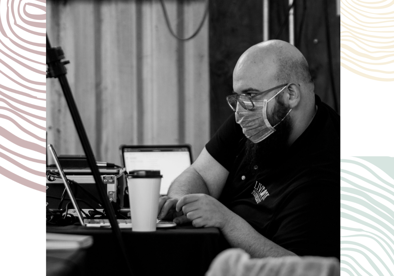 A person wearing glasses and a face mask sits at a table working on a laptop, surrounded by audio-visual equipment, with a coffee cup nearby.