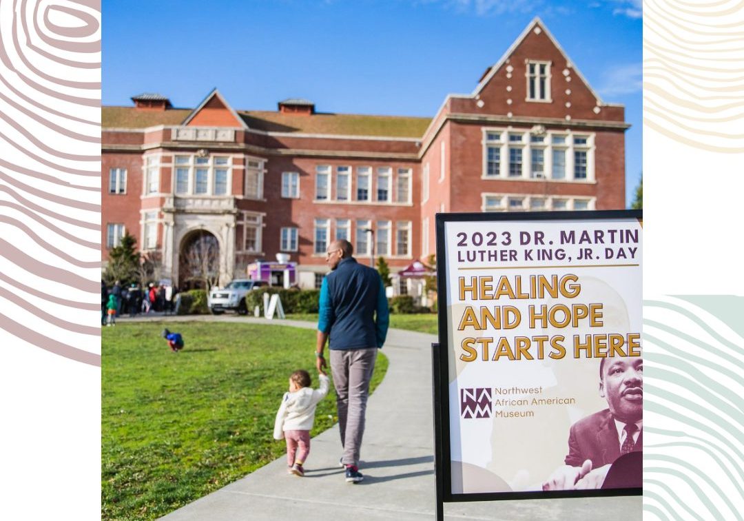 A man and a child walk toward a brick building; a sign in the foreground promotes the 2023 Dr. Martin Luther King, Jr. Day virtual keynote with Jesse Williams at the Northwest African American Museum.