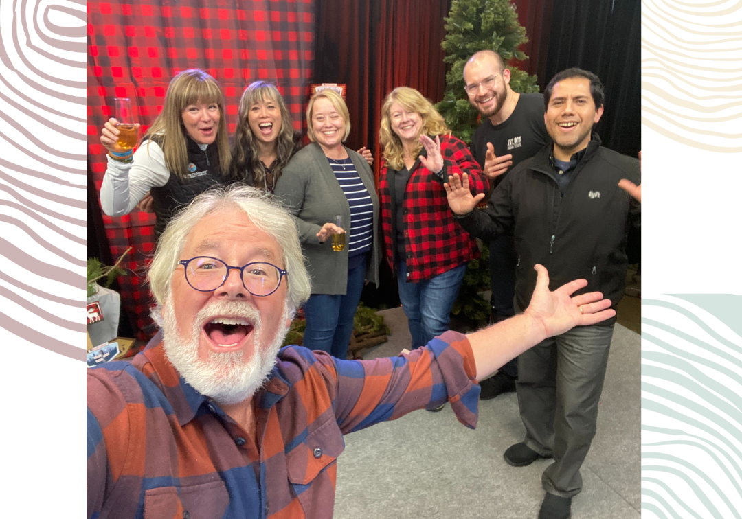 A man with white hair and beard stands with his arm outstretched to a group of six people behind him.