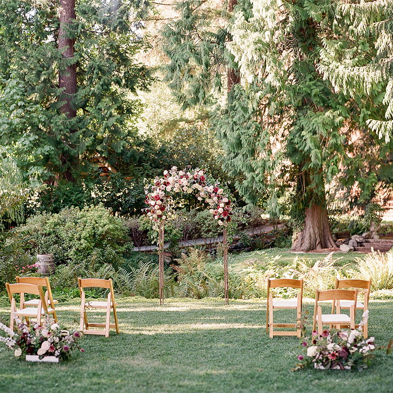 PC-Emily-Ganey_Jing-David-Wedding-224 A small outdoor wedding ceremony setup with wooden chairs facing a floral arch, surrounded by lush greenery and tall trees—perfect for Seattle weddings and private events that may need AV services.