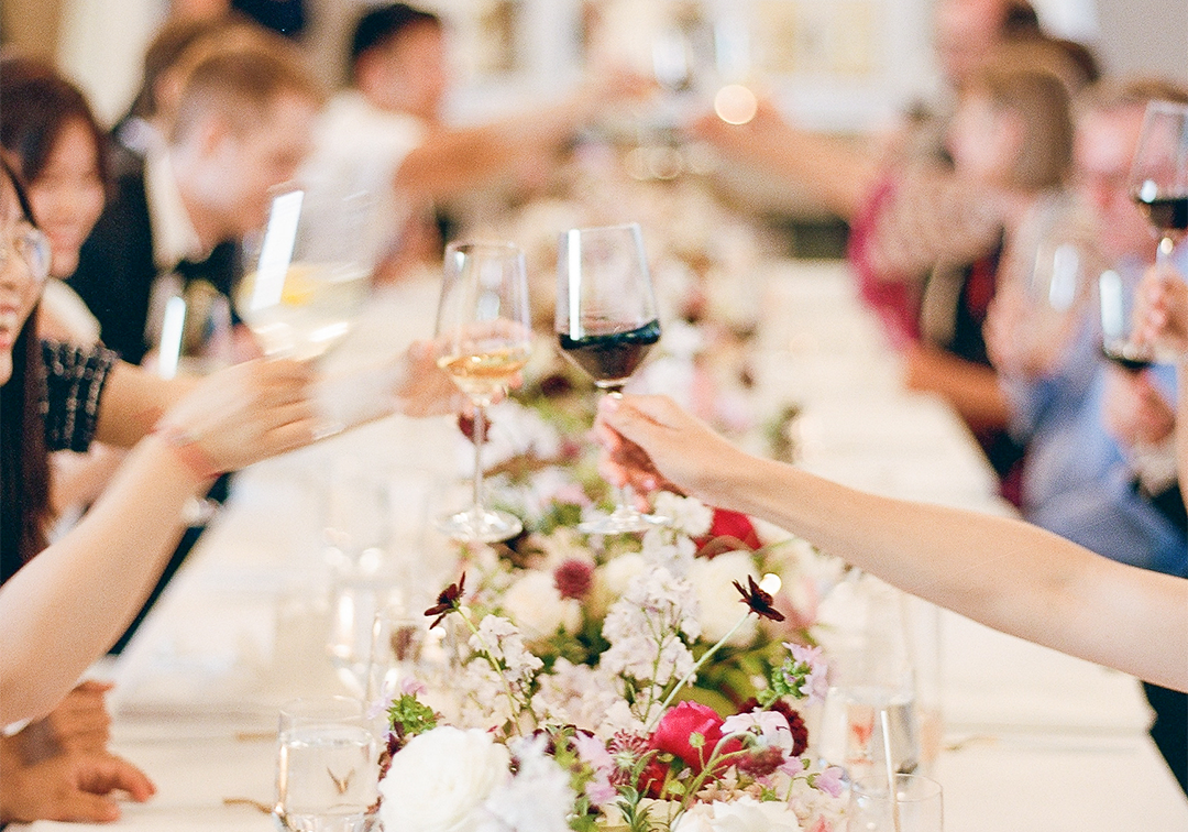 People seated at a long table raise their wine glasses in a toast, with floral arrangements running down the center—perfect for Seattle weddings and private events enhanced by professional AV services.