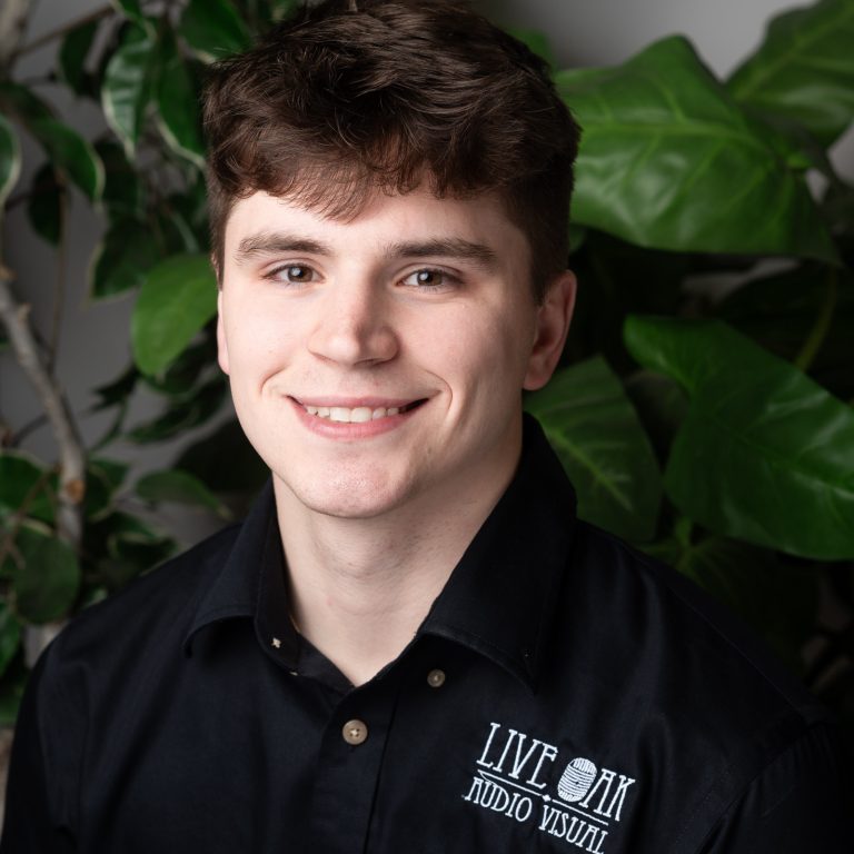 Paul Pease HS 2024 A young man with brown hair, wearing a black "Live On Audio Visual" shirt, smiles at the camera with green leafy plants in the background—proudly representing the Above-and-Beyond Client Experience Team.