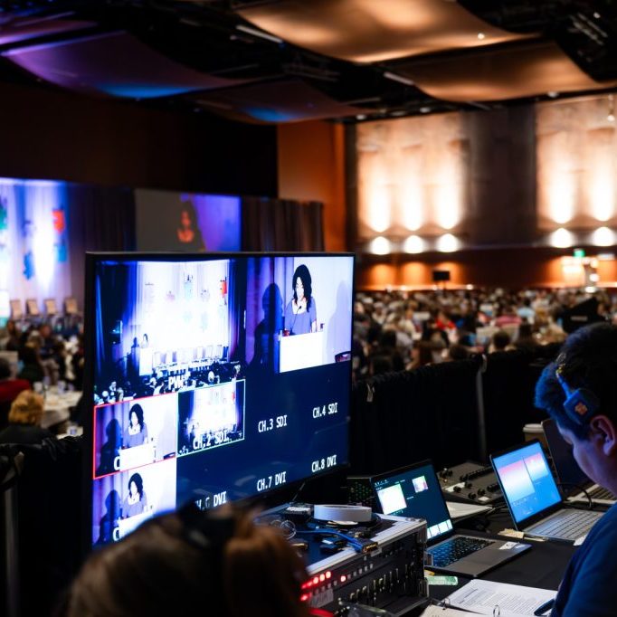 A technician with Production Partners operates audio-visual equipment at the back of a large conference room, with multiple screens showing a speaker addressing an audience.