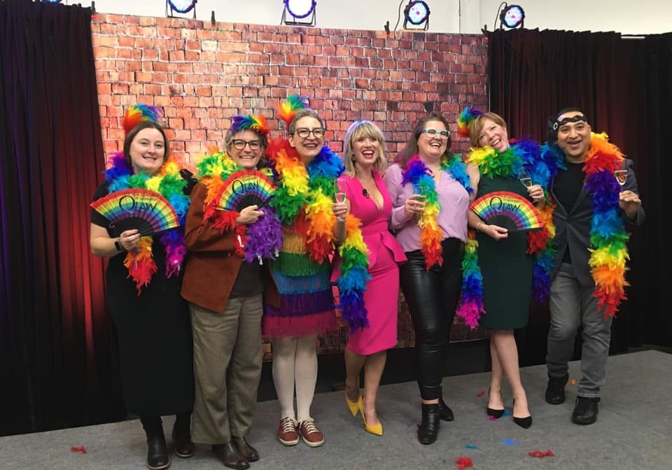 Six people stand in front of a brick wall backdrop at the Falltacular event, wearing colorful rainbow feather boas and holding rainbow fans, smiling for the camera in support of the QLaw Foundation of Washington.