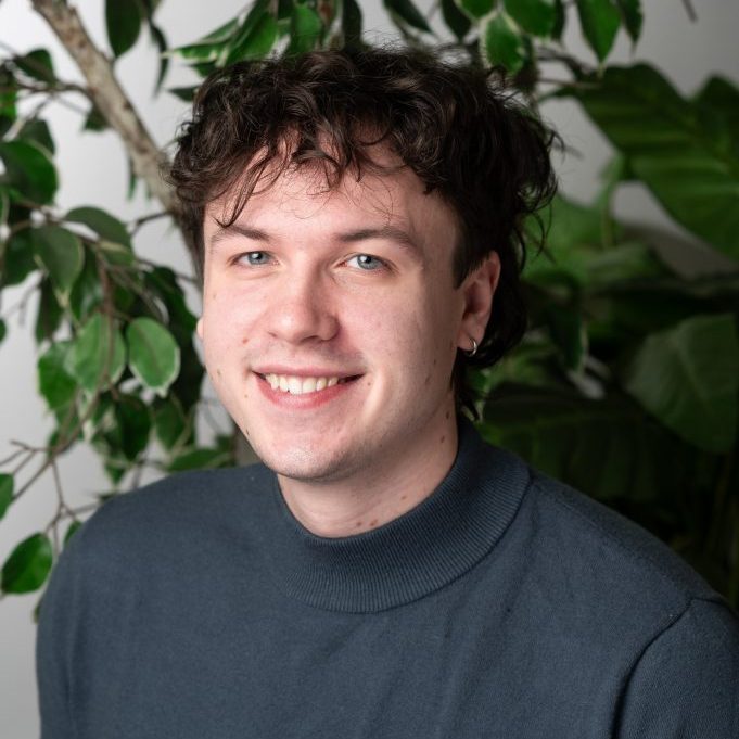 Sam Yates 2024 A young man with wavy brown hair and a blue sweater smiles at the camera, representing the Above-and-Beyond Client Experience Team while sitting in front of green leafy plants.