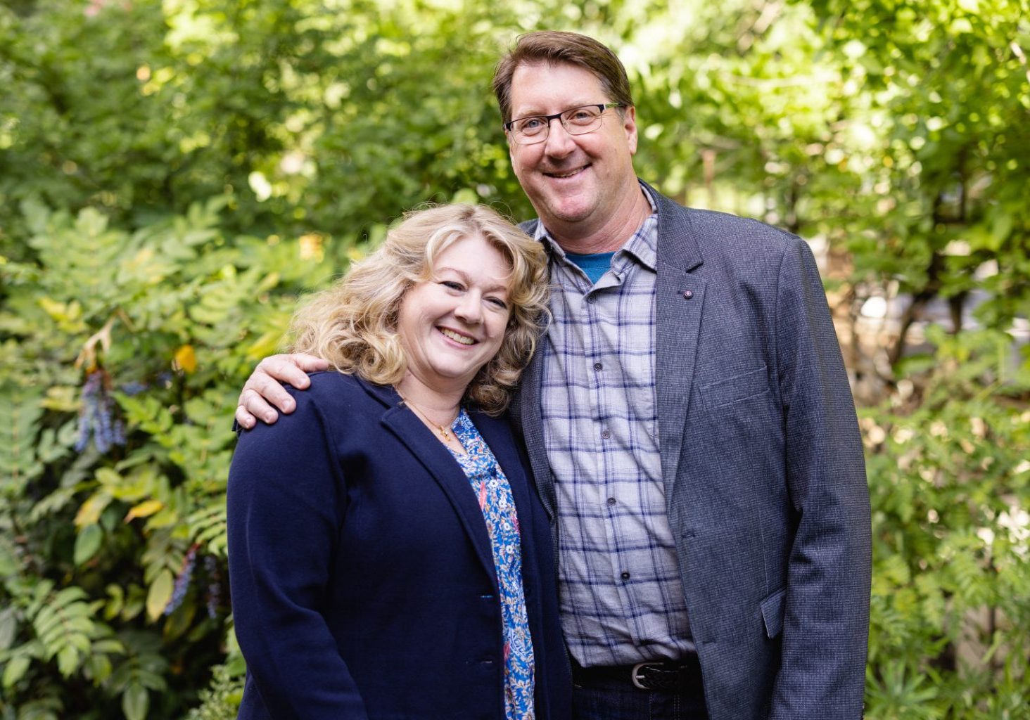 A woman and man stand close together outdoors, smiling at the camera, with greenery in the background.
