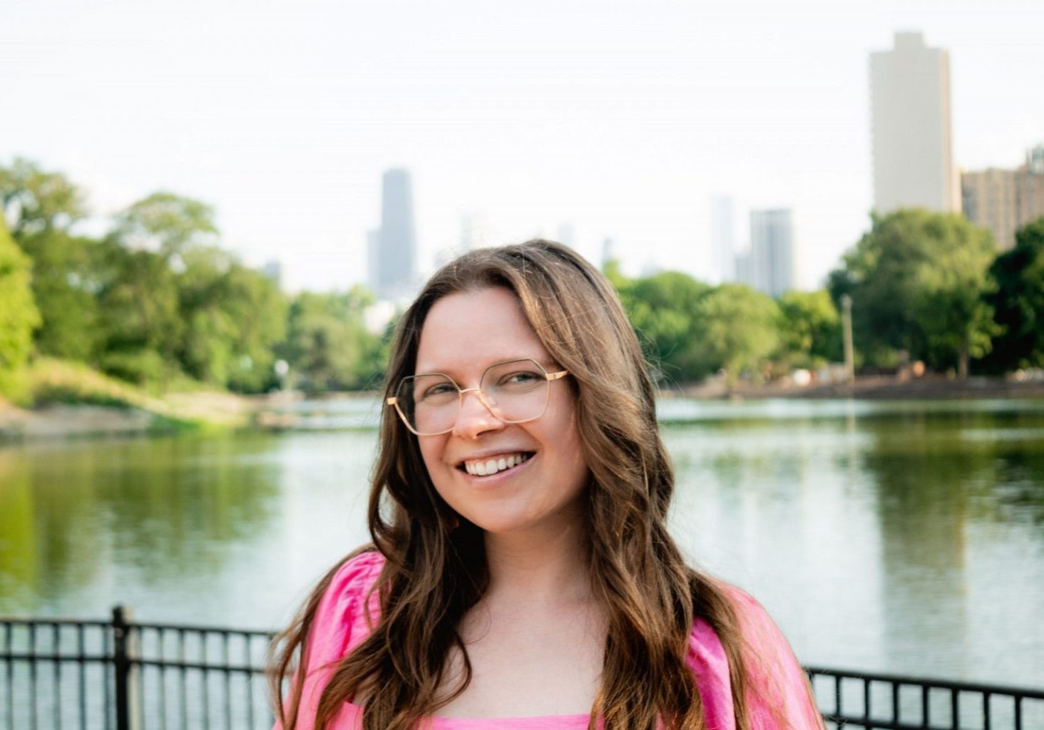 A woman with long brown hair and glasses smiles in front of a pond with trees and city buildings in the background on a sunny day, breaking misconceptions about working in corporate AV.