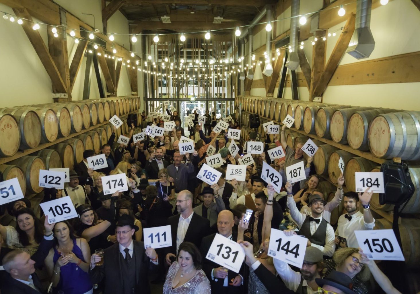 A large group at an indoor event hold numbered signs, surrounded by wine barrels and string lights in a rustic setting—perfect for hybrid events that combine in-person energy with virtual connections.