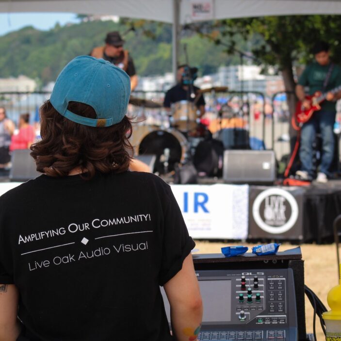 Photo of the back of a Live Oak AV employee as they adjust the sound for live music at an event. Their shirt says "Amplifying Our Community. Live Oak Audio Visual".