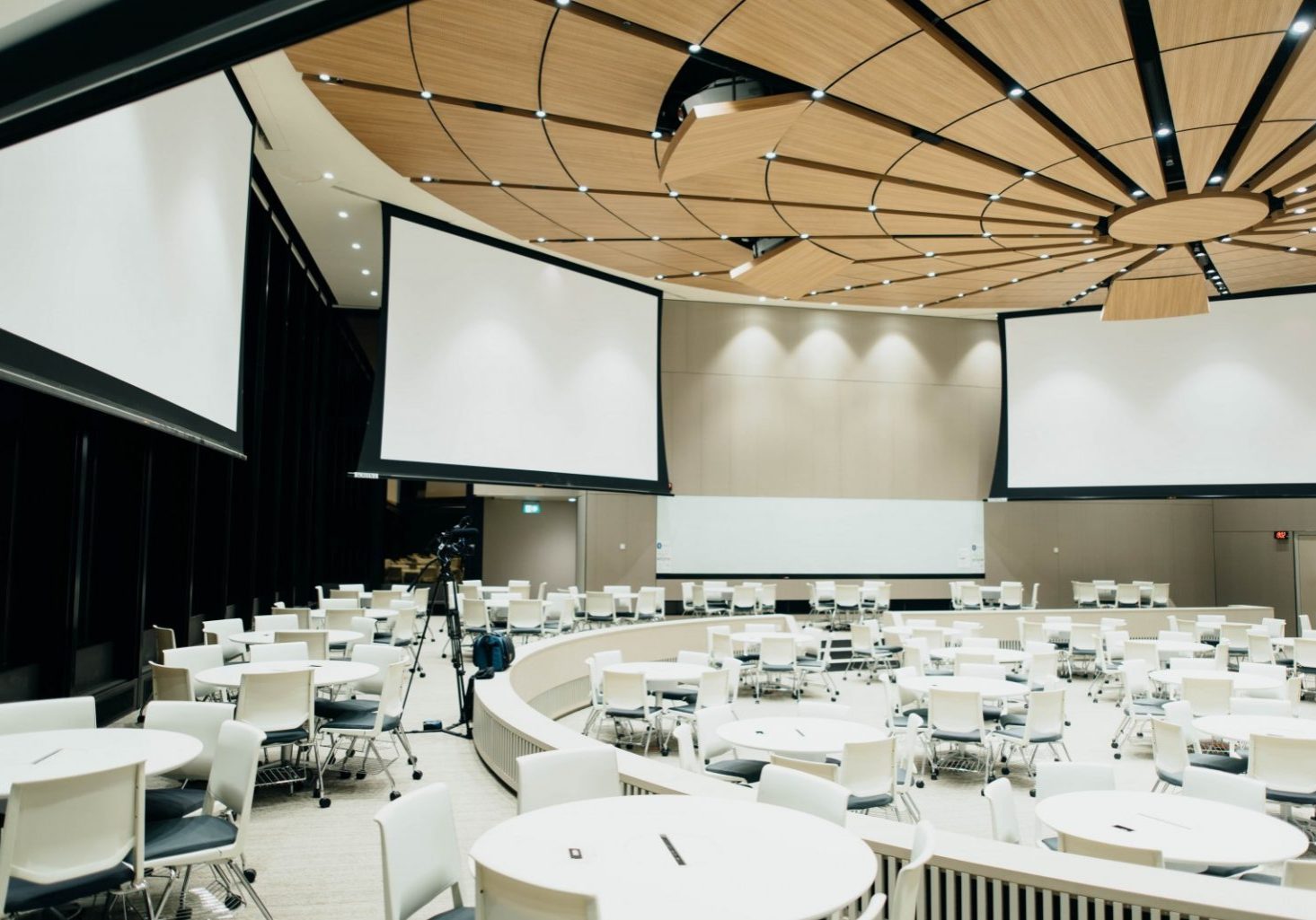Large modern conference room with round tables, white chairs, several projection screens, a whiteboard, and a camera on a tripod—ideal for hybrid events and seamless event planning. The ceiling features an elegant wood panel design.
