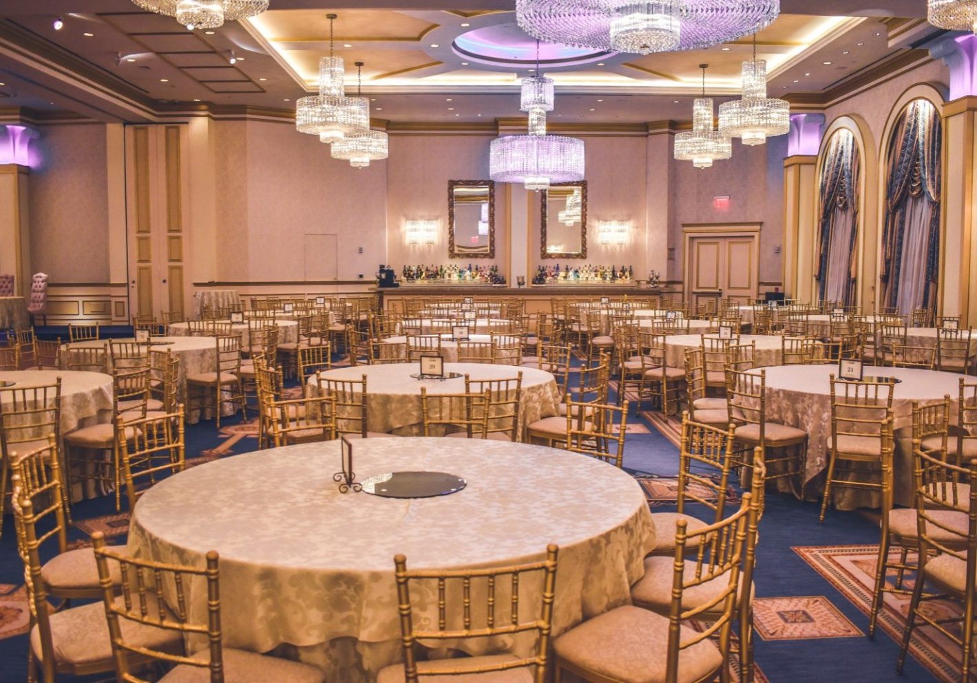 Spacious banquet hall with round tables in cream, gold chairs, ornate chandeliers, and a bar area—perfect for event planning or hosting fall events. The empty room is well-lit, ready to accommodate any occasion amid uncertainty.
