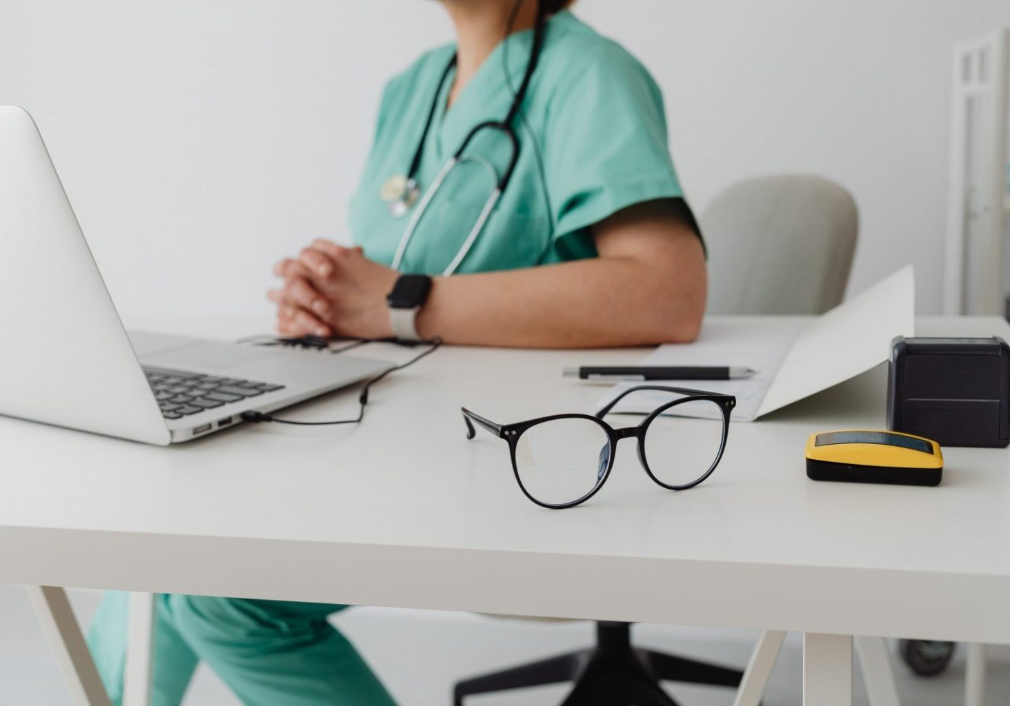 A healthcare professional in green scrubs sits at a desk with a laptop, folded hands, eyeglasses, and medical equipment visible in the foreground, preparing for a live streaming Continuing Medical Education session.
